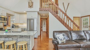A spacious kitchen with a central island, wooden bar stools, a brown leather couch, and a wooden staircase.