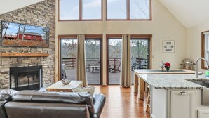 A spacious living room with a stone fireplace, leather sofa, and a large window overlooking a deck.