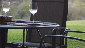 Una mesa al aire libre con dos copas de vino blanco, un pan y un cuchillo sobre una superficie metálica y sillas.