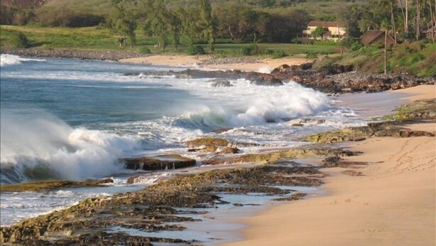 Una playa costera con costa rocosa, arena y casas al fondo.