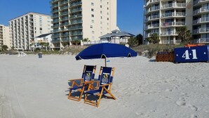 Ein Strand mit zwei Liegestühlen unter einem blauen Sonnenschirm, einem Holzzaun und einem Gebäude mit Balkonen.