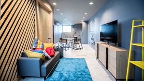 A modern living room with a sofa, colorful cushions, a blue rug, a wooden accent wall, a dining area with chairs, a TV, and a yellow ladder. - The CAPSULE by UnionSPACE - Adults Only (Bangkok)