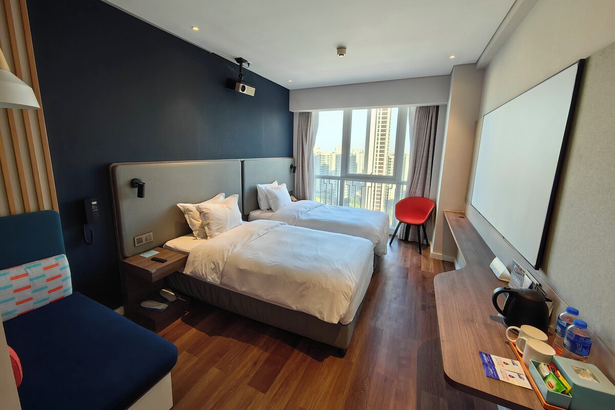 A hotel room with two beds, a desk with a kettle, a red chair, and a large window with a city view.