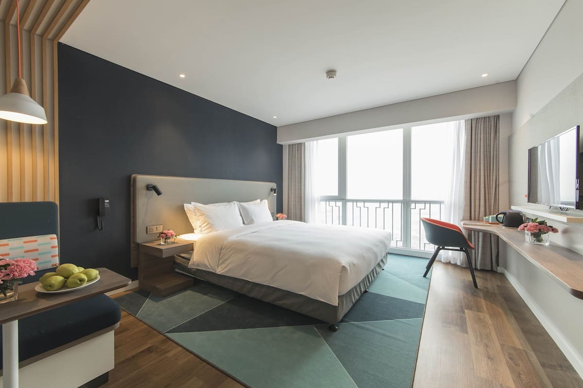 Ein modernes Hotelzimmer mit einem großen Bett, einem Nachttisch und einem Schreibtisch mit Stuhl. Ein Fenster mit Vorhängen und ein Fernseher an der Wand sind ebenfalls vorhanden.