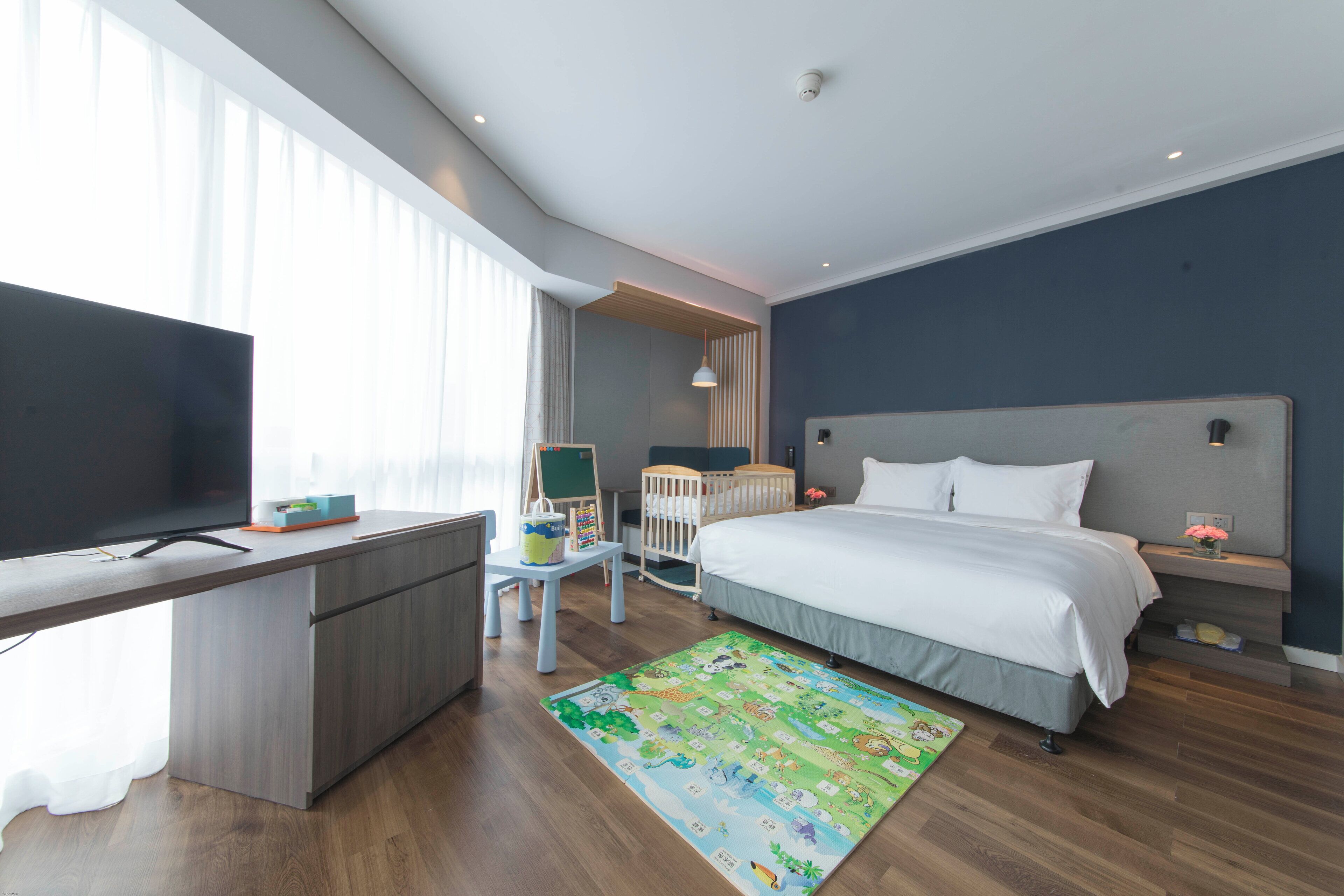 A modern hotel room with a wooden desk, a flat-screen TV, a bed with white bedding, a colorful rug, and a crib.