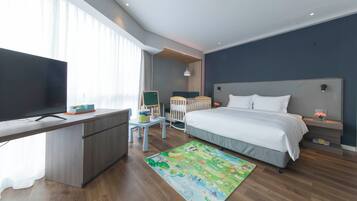 A modern hotel room with a wooden desk, a flat-screen TV, a bed with white bedding, a colorful rug, and a crib.