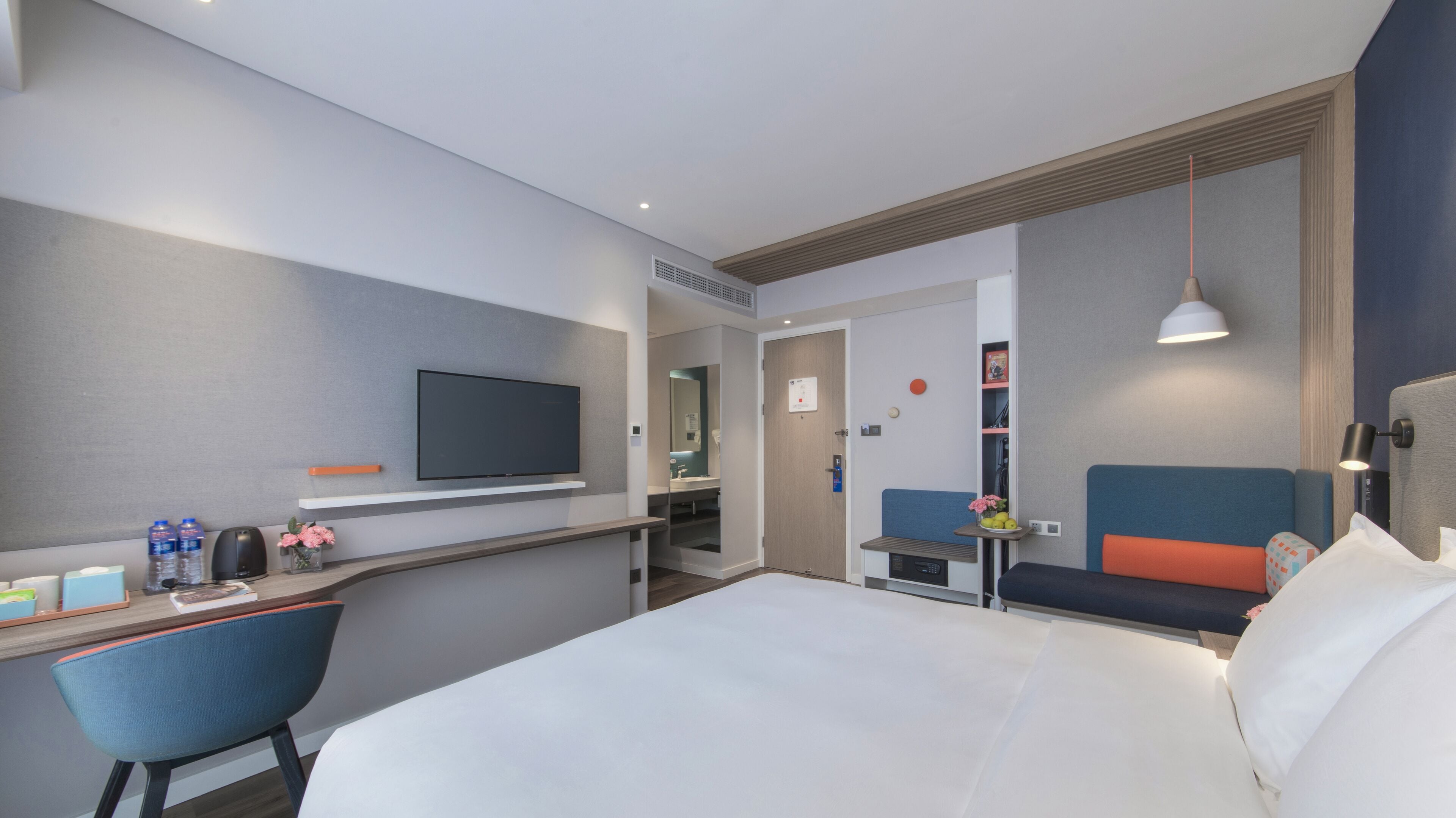 A modern hotel room with a bed, a desk with a flat-screen TV, a chair, a small table with a vase of flowers, and a sofa.