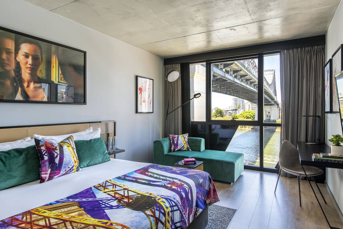 Une chambre d’hôtel moderne dotée d’un grand lit, d’un canapé vert, d’un bureau avec une chaise, et offrant une vue sur un pont et le paysage urbain à travers de grandes fenêtres.