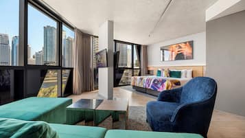 A modern hotel room with a large window, a bed with colorful bedding, a blue armchair, and a small table.