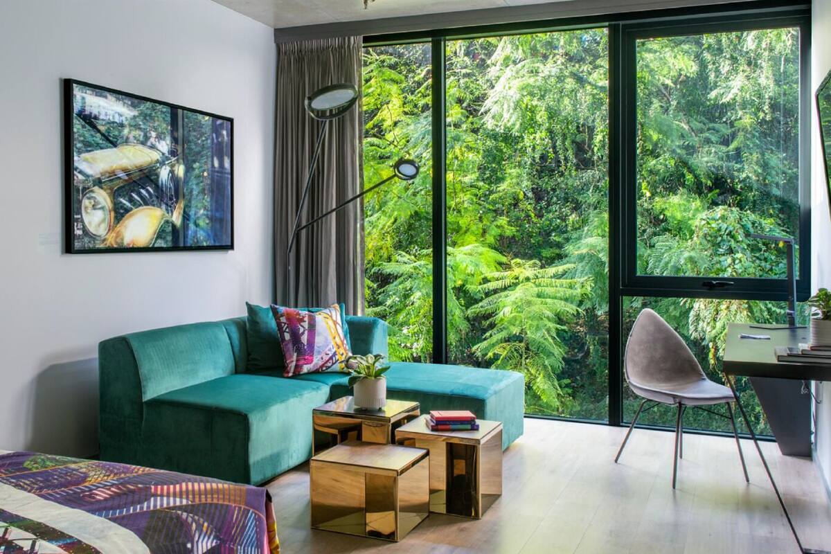 Une chambre moderne avec un canapé de couleur turquoise, une grande fenêtre donnant sur une forêt, un petit bureau avec une chaise et un tableau accroché au mur.