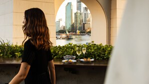 A woman standing by a window overlooking a cityscape with buildings and a river. - Crystalbrook Vincent (Brisbane)