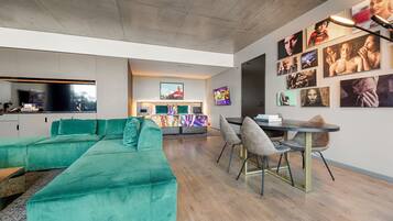 A modern living room with a green sectional sofa, a dining area with a round table and chairs, and a wall decorated with various framed pictures.