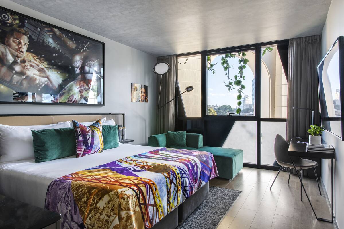 Une chambre d’hôtel moderne dotée d’un grand lit, de sièges confortables recouverts d’un tissu vert, d’un bureau et offrant une vue sur la ville grâce à de grandes fenêtres.