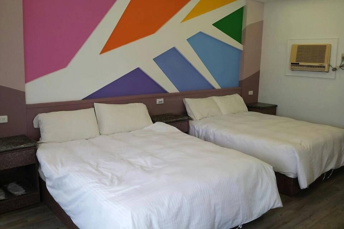A hotel room with two beds, a colorful geometric wall design, and a small window with an air conditioning unit.