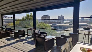 Ein moderner Außenbereich zum Sitzen mit Blick auf die Stadt, ausgestattet mit verschiedenen Stühlen und Tischen und einer transparenten Glasabtrennung.