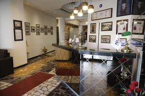 Un hall d’entrée avec un comptoir de réception, des tableaux encadrés aux murs, un sol à motifs et un escalier en arrière-plan.