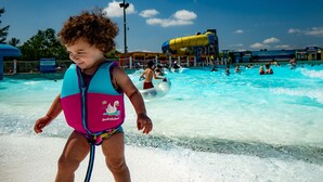 Criança com colete de natação em parque aquático, com um toboágua e outras pessoas ao fundo.