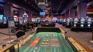 Uma mesa de craps em um cassino, com várias máquinas caça-níqueis e uma tela grande ao fundo.
