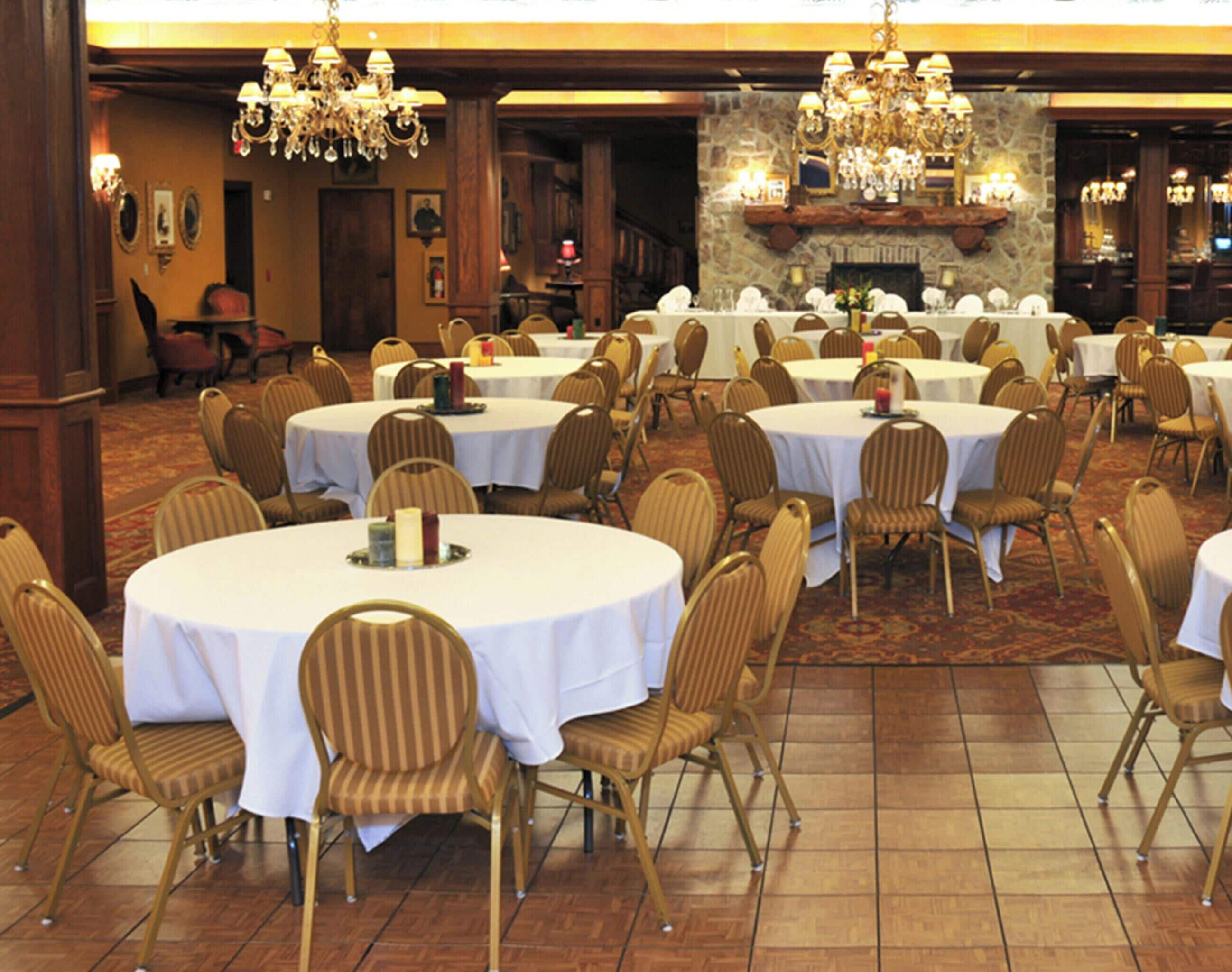 A banquet hall with round tables and chairs, chandeliers, and a stone fireplace.