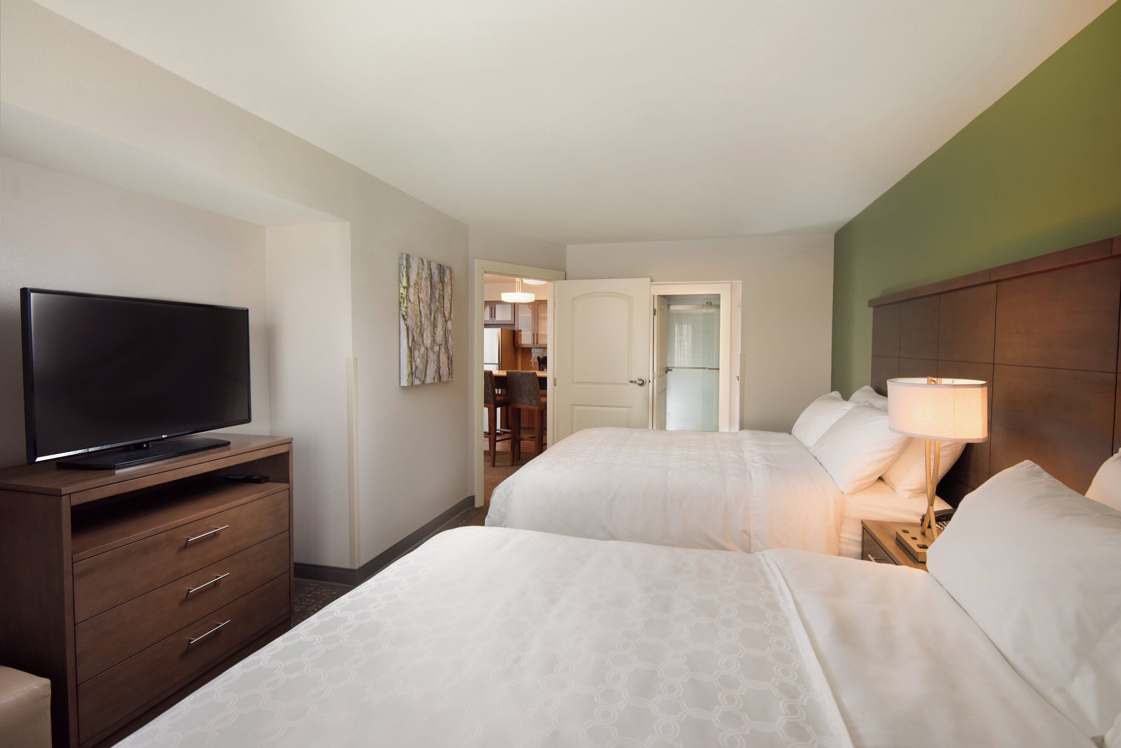 Habitación de hotel con una televisión sobre una cómoda de madera, dos camas con ropa blanca, una mesita de noche con una lámpara y un pasillo que conduce a un baño.