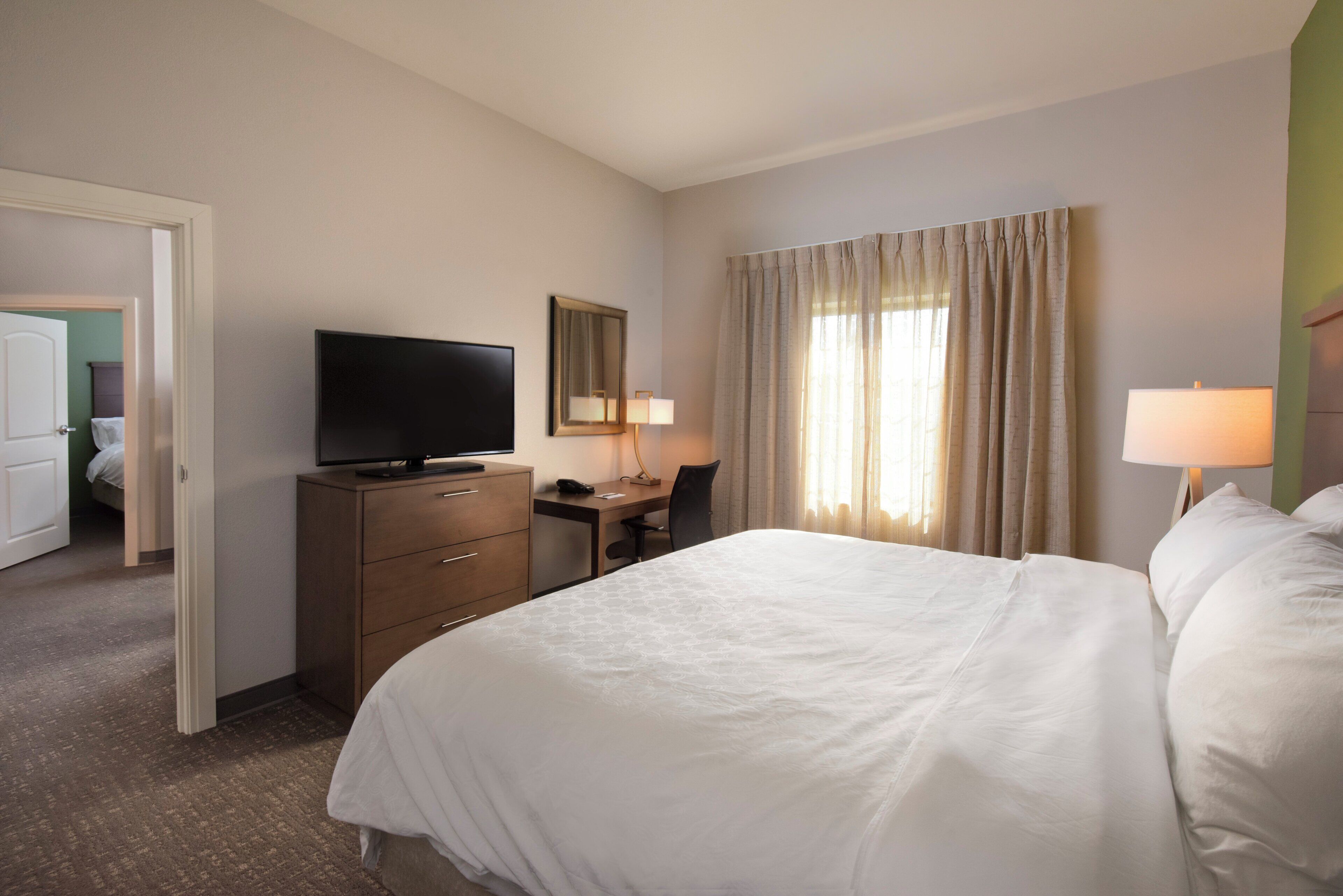 Een hotelkamer met een bed, een bureau, een televisie, een stoel, een lamp en een deur naar een andere kamer.