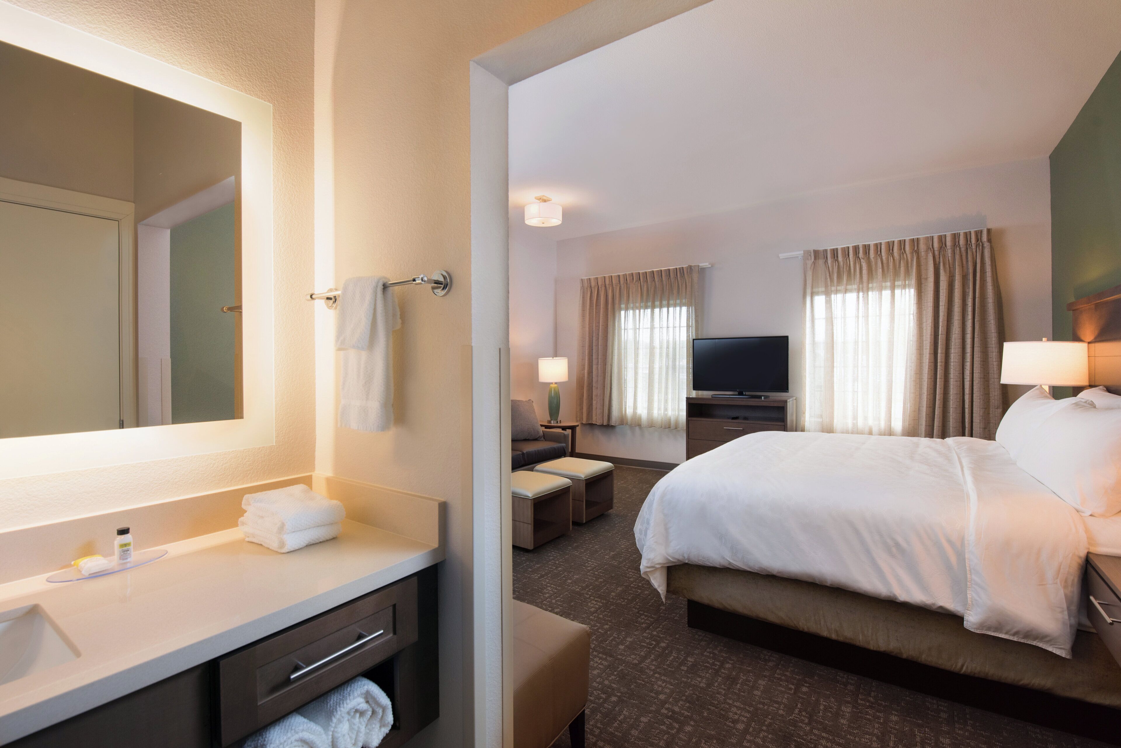 Une chambre d’hôtel dotée d’un grand lit, d’un coin salon avec un canapé et des fauteuils, d’une télévision à écran plat, d’une salle de bain avec un miroir et d’une coiffeuse avec un lavabo et un porte-serviettes.