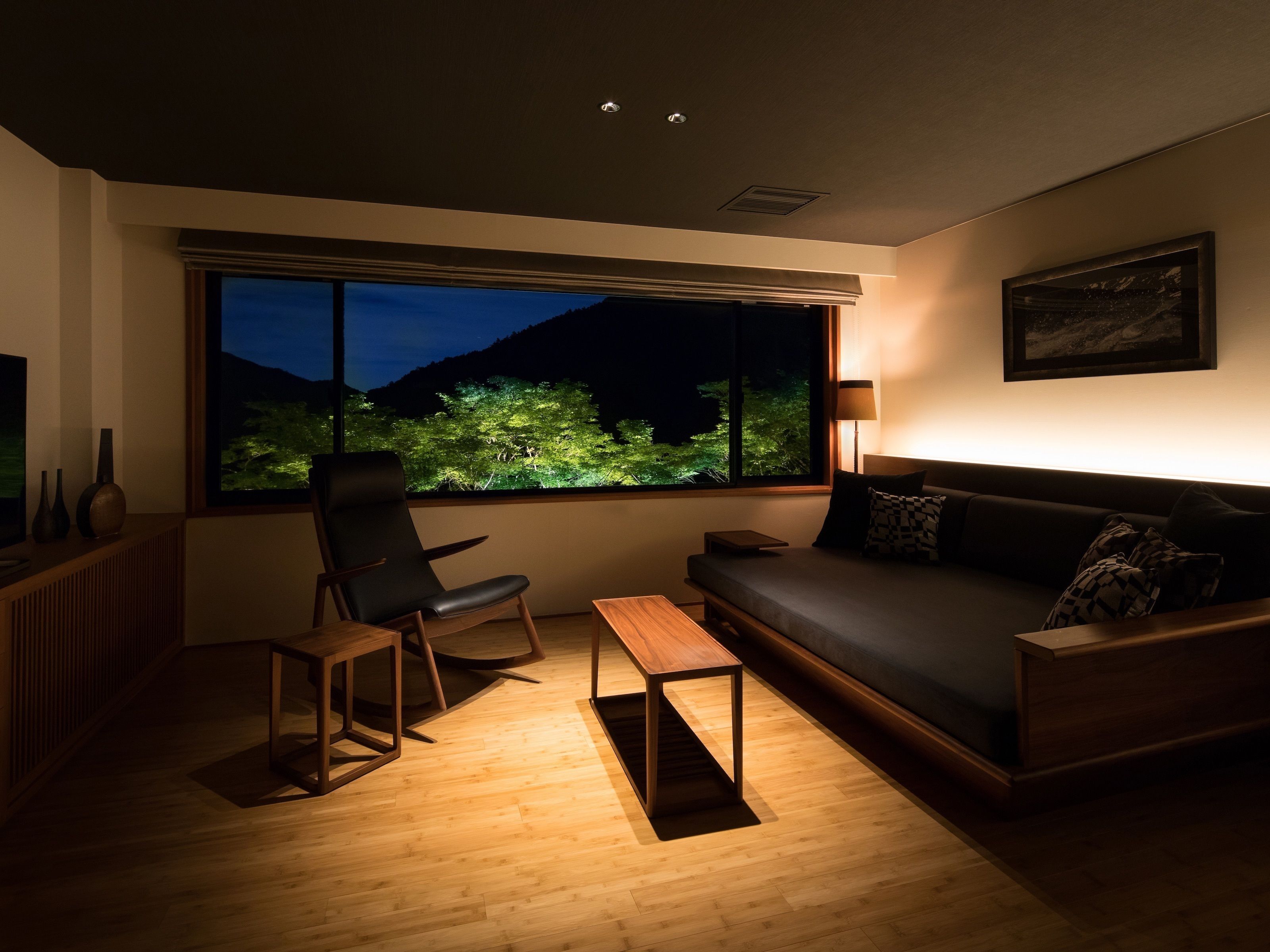 A modern living room with a large window, a sofa, a chair, a small table, and a lamp.