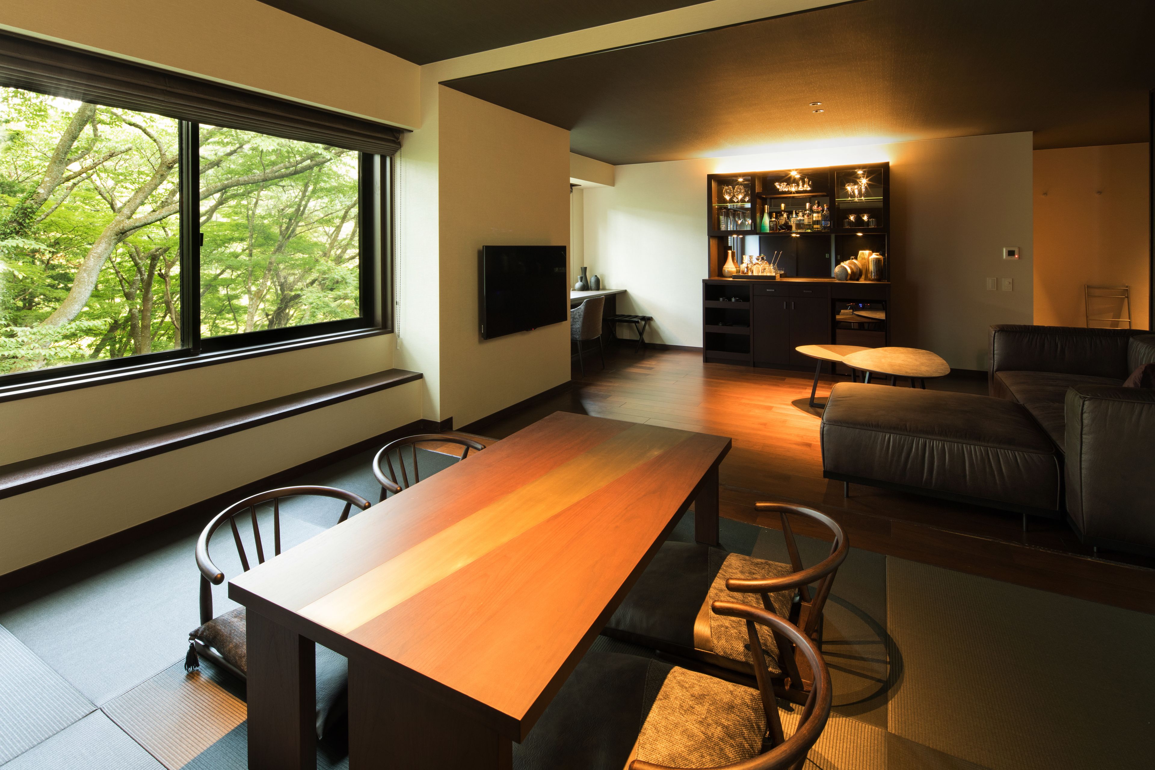 A modern living room with a wooden dining table, leather sofa, and a kitchen area with bar shelves.