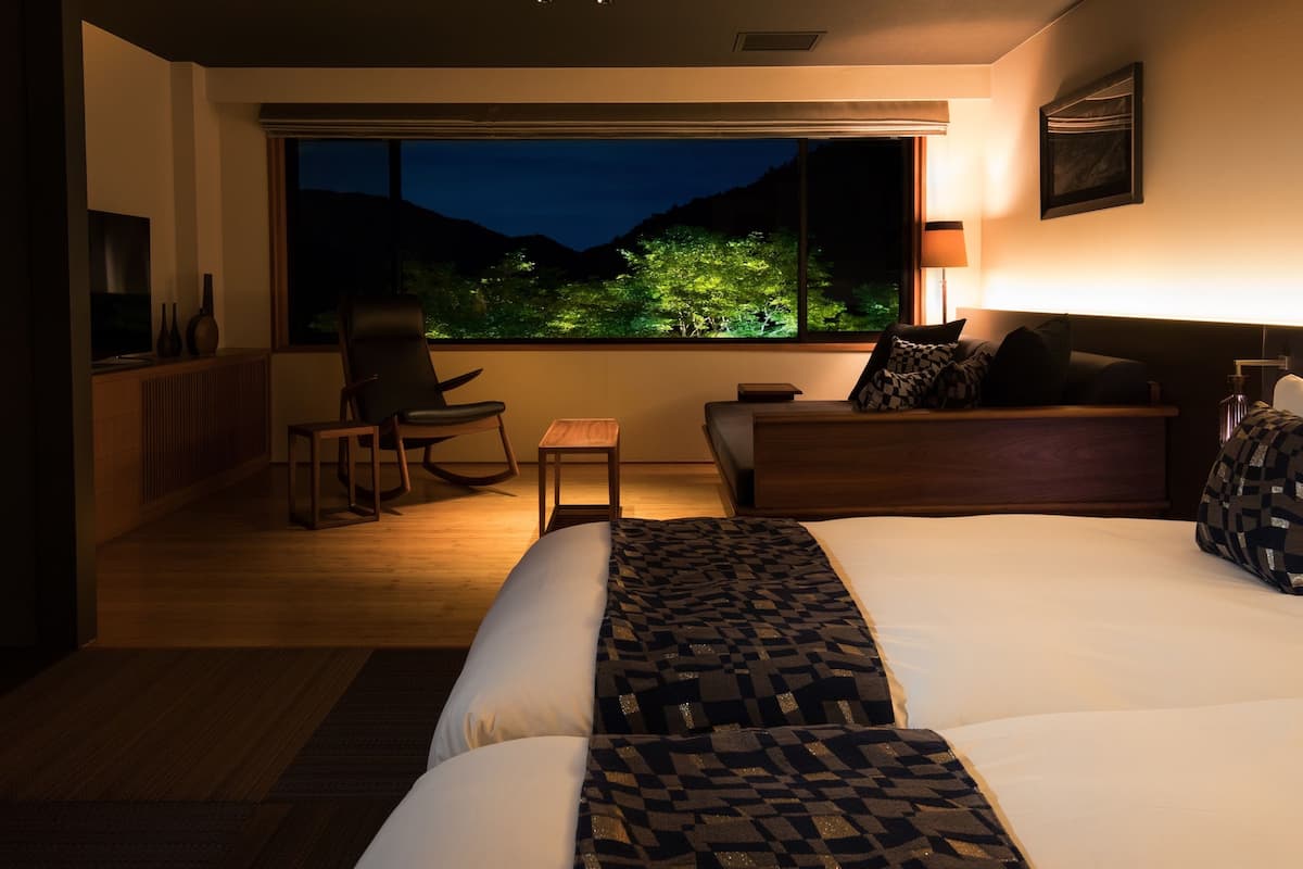 Habitación de hotel con una cama grande, zona de estar con una silla y una mesita, un sofá y vistas a un cielo nocturno con árboles.