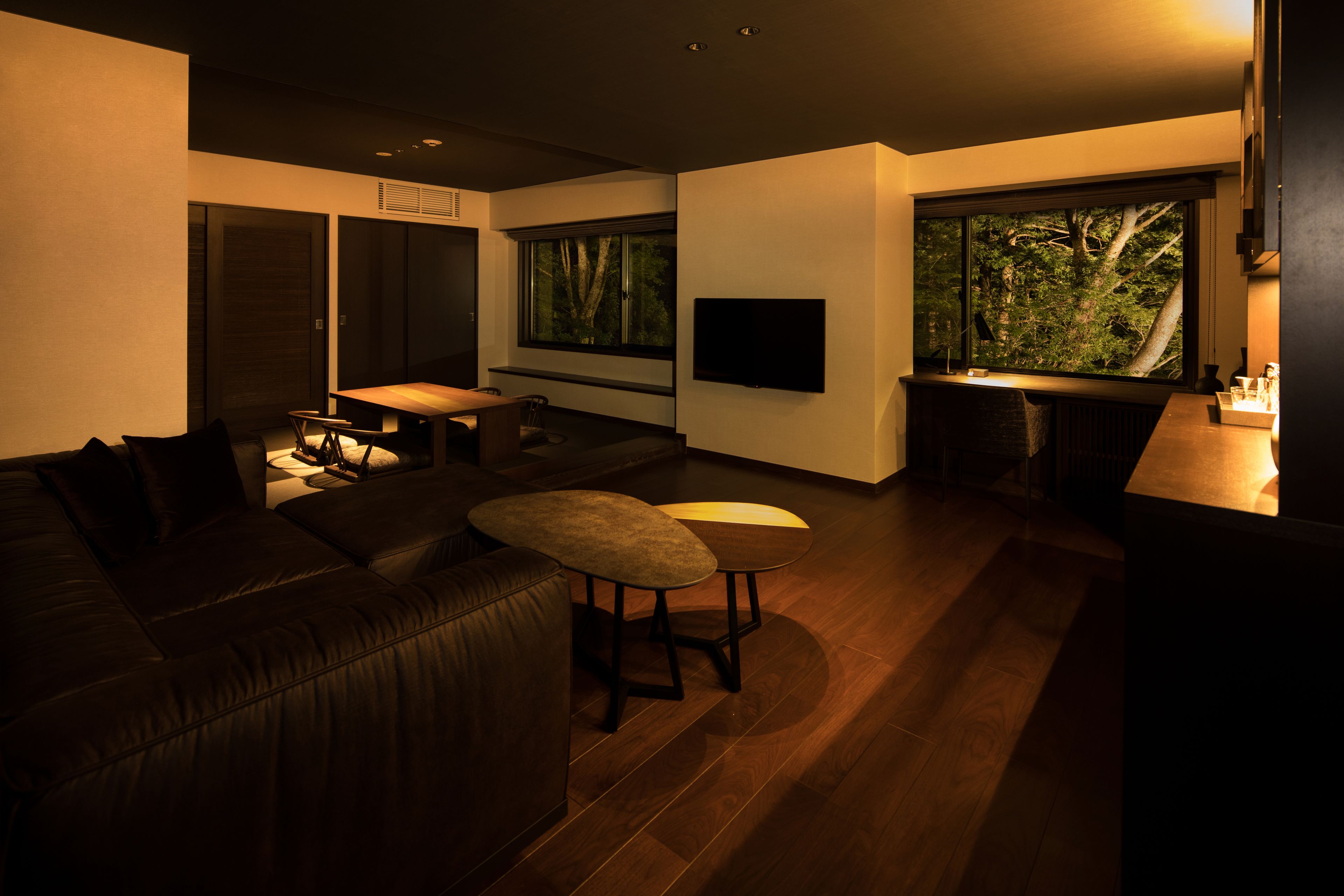 A modern living room with a dark leather sofa, a small round table, and a large window with a view of trees.