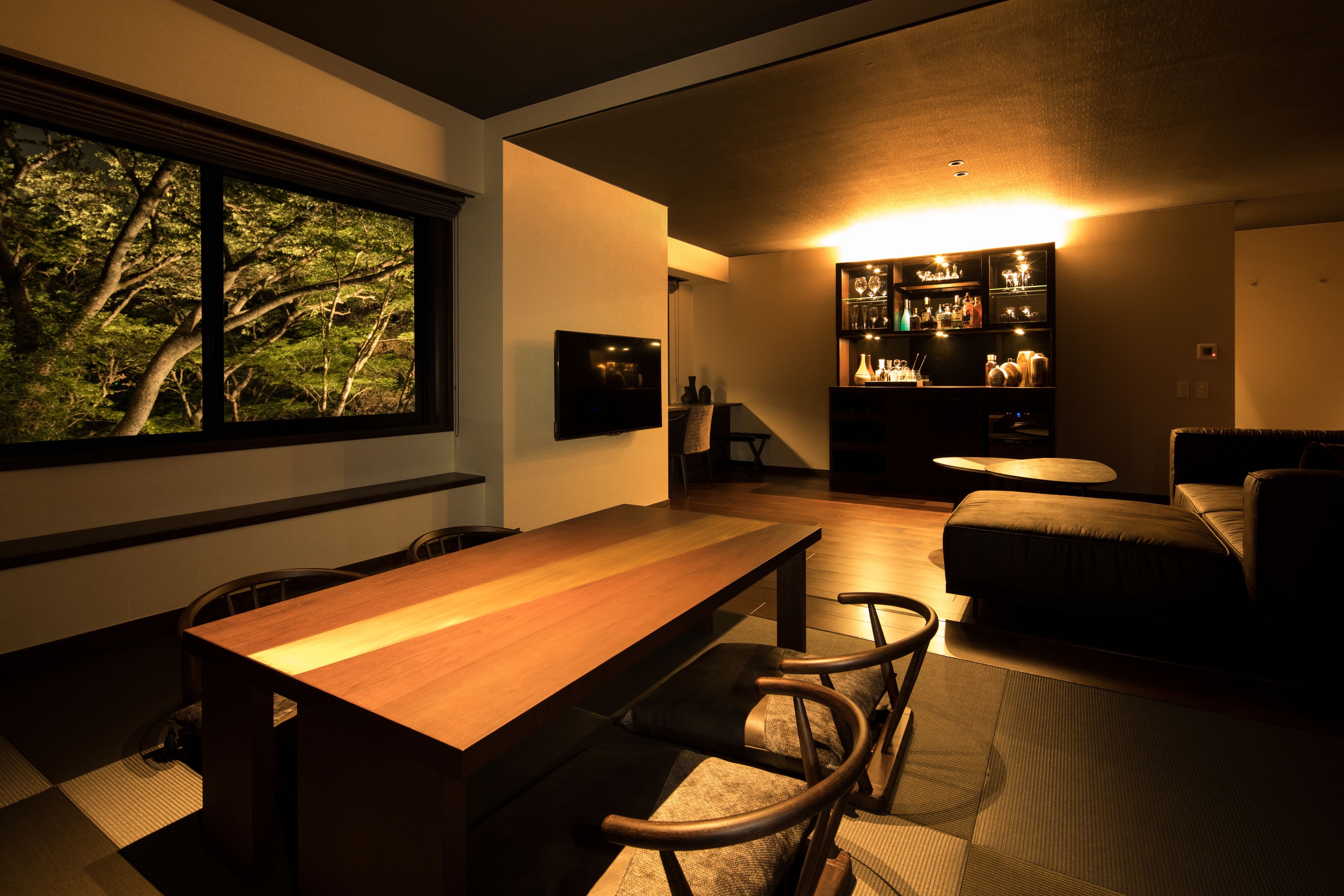 A modern living room with a wooden dining table, a sofa, and a coffee table. There is a large window with a view of trees.