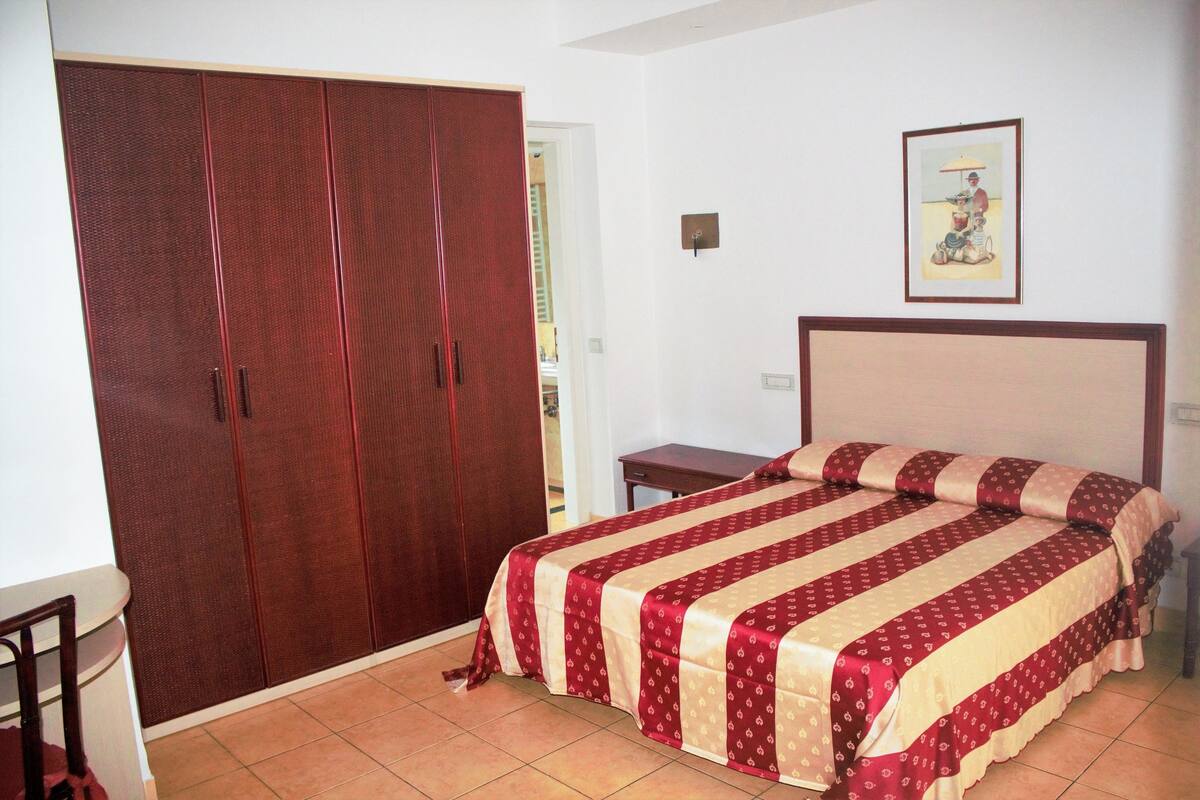Una camera da letto con un ampio armadio, un letto con copriletto a righe, un comodino e una lampada a parete.