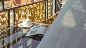 Een balkon met uitzicht op strand en stadsbeeld, met een tafel, een beker, een rieten stoel en een bed met een wit laken.