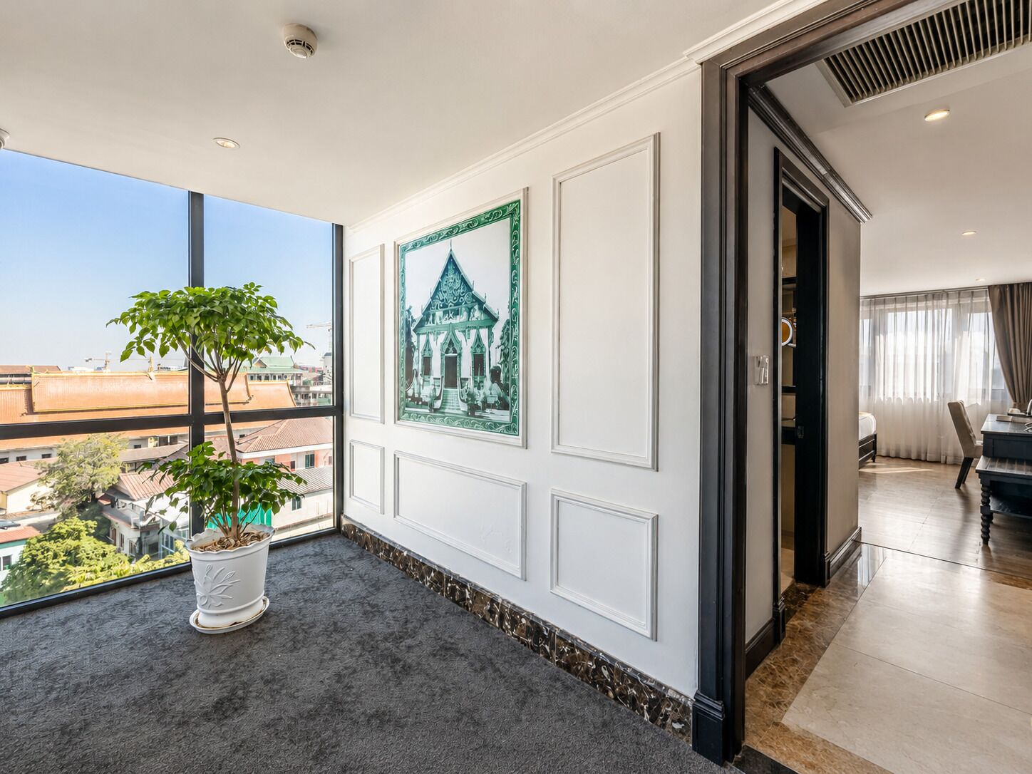 A hotel room with a large window, a potted plant, a framed picture, and a view of the cityscape.