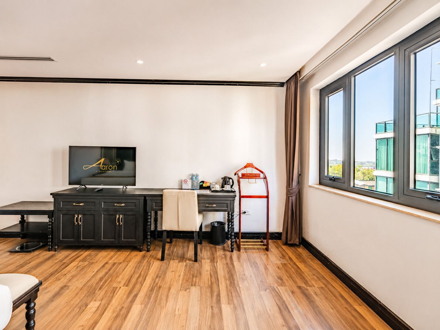 Habitación de hotel con piso de madera, un escritorio con televisor, una silla, un perchero rojo y una ventana con vista a edificios.