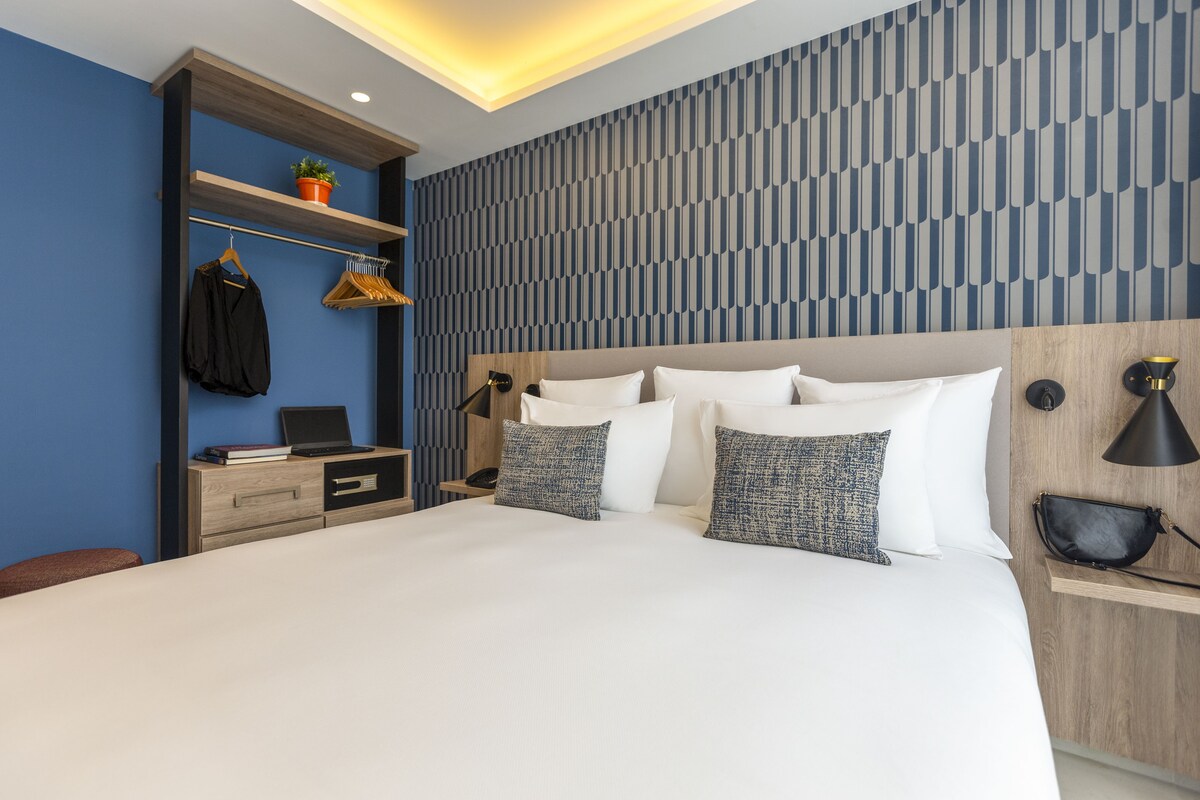 A modern hotel room with a large bed, a bedside table, a desk with a laptop, a shelf with a plant, and a patterned wall.