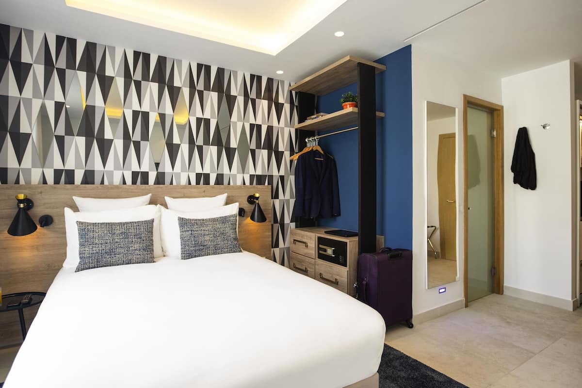 A modern hotel room with a large bed, a wardrobe, a desk, and a mirror.