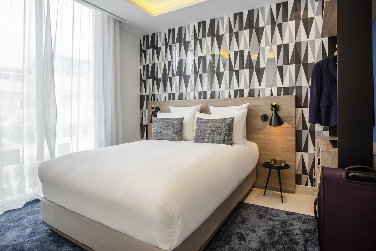 A modern hotel room with a large bed, a geometric-patterned wall, a small bedside table, and a coat rack.