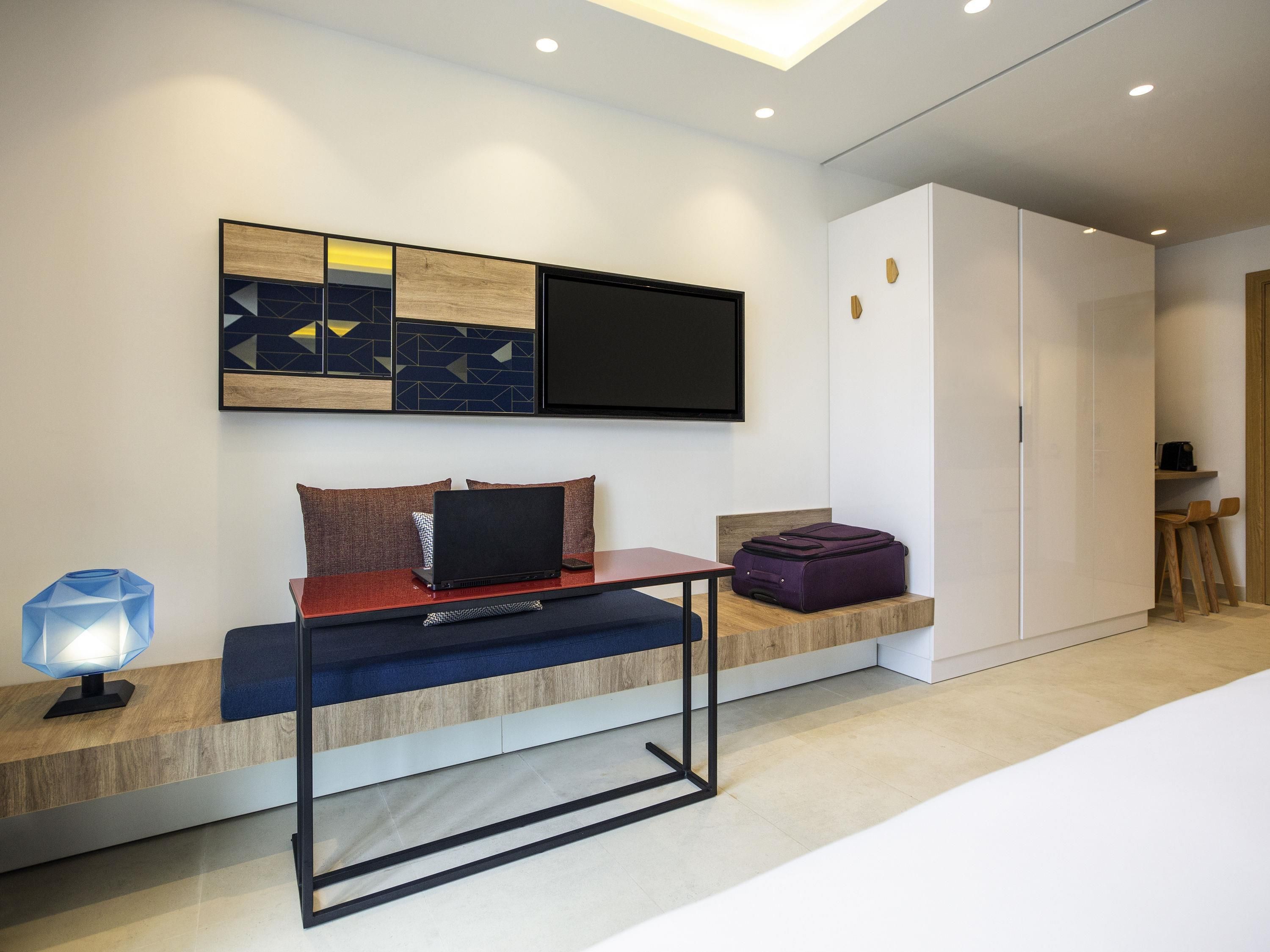 Una habitación de hotel moderna con un televisor de pantalla plana montado en la pared, un ordenador portátil sobre un banco de madera, una maleta morada y un escritorio con una lámpara.