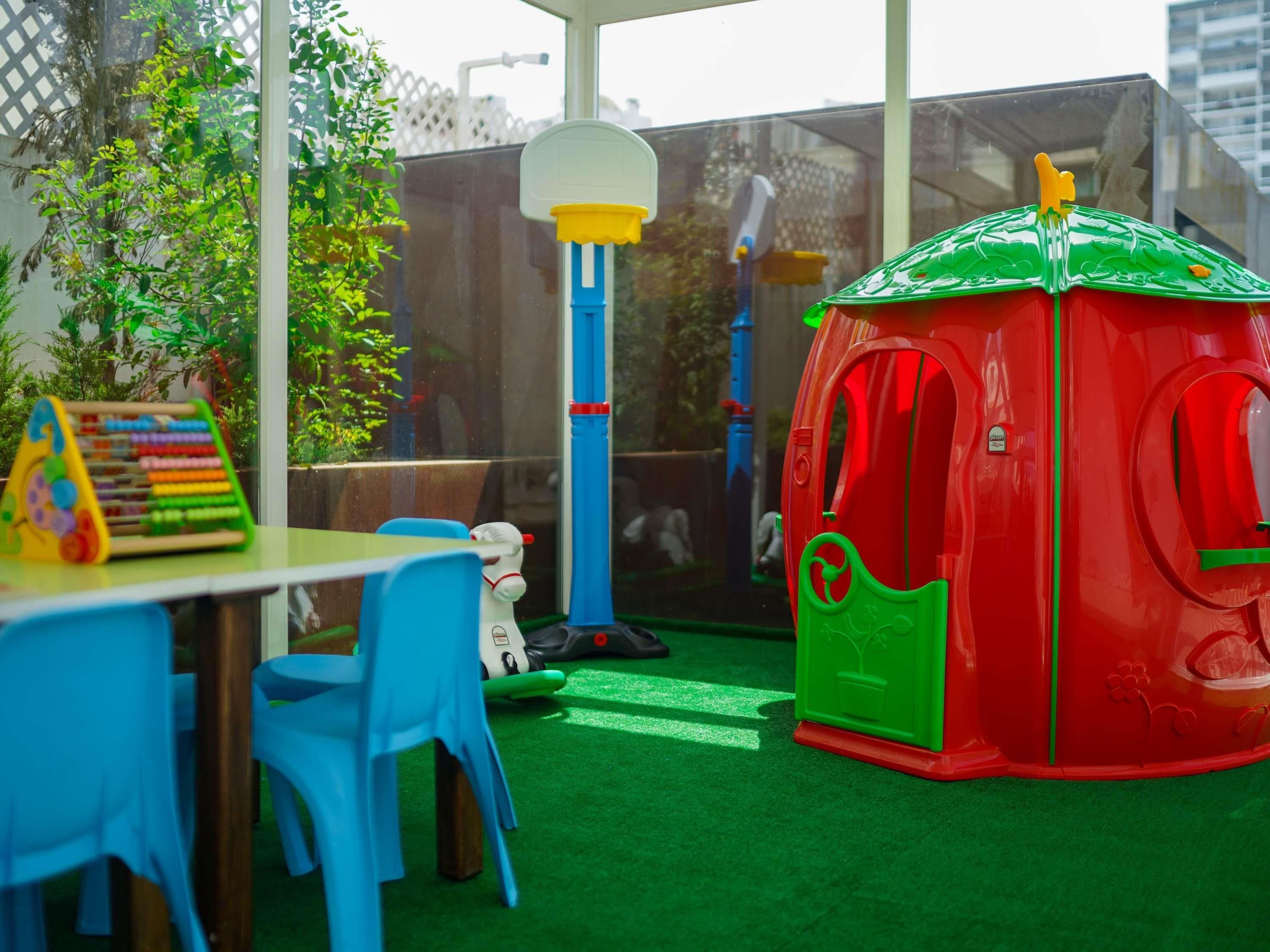 Un espace de jeux pour enfants avec une maisonnette rouge, un panier de basket et une table à jeux sur laquelle est posé un abaque coloré.