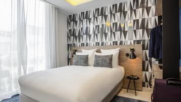 A modern hotel room with a large bed, a geometric-patterned wall, a small bedside table, and a coat rack.