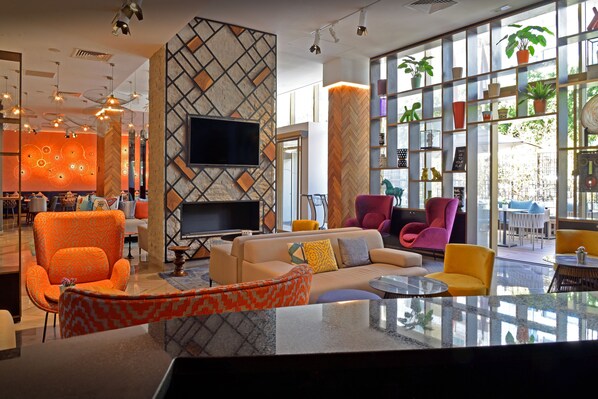 A modern hotel lobby with a large TV, comfortable seating, and a large window with plants.