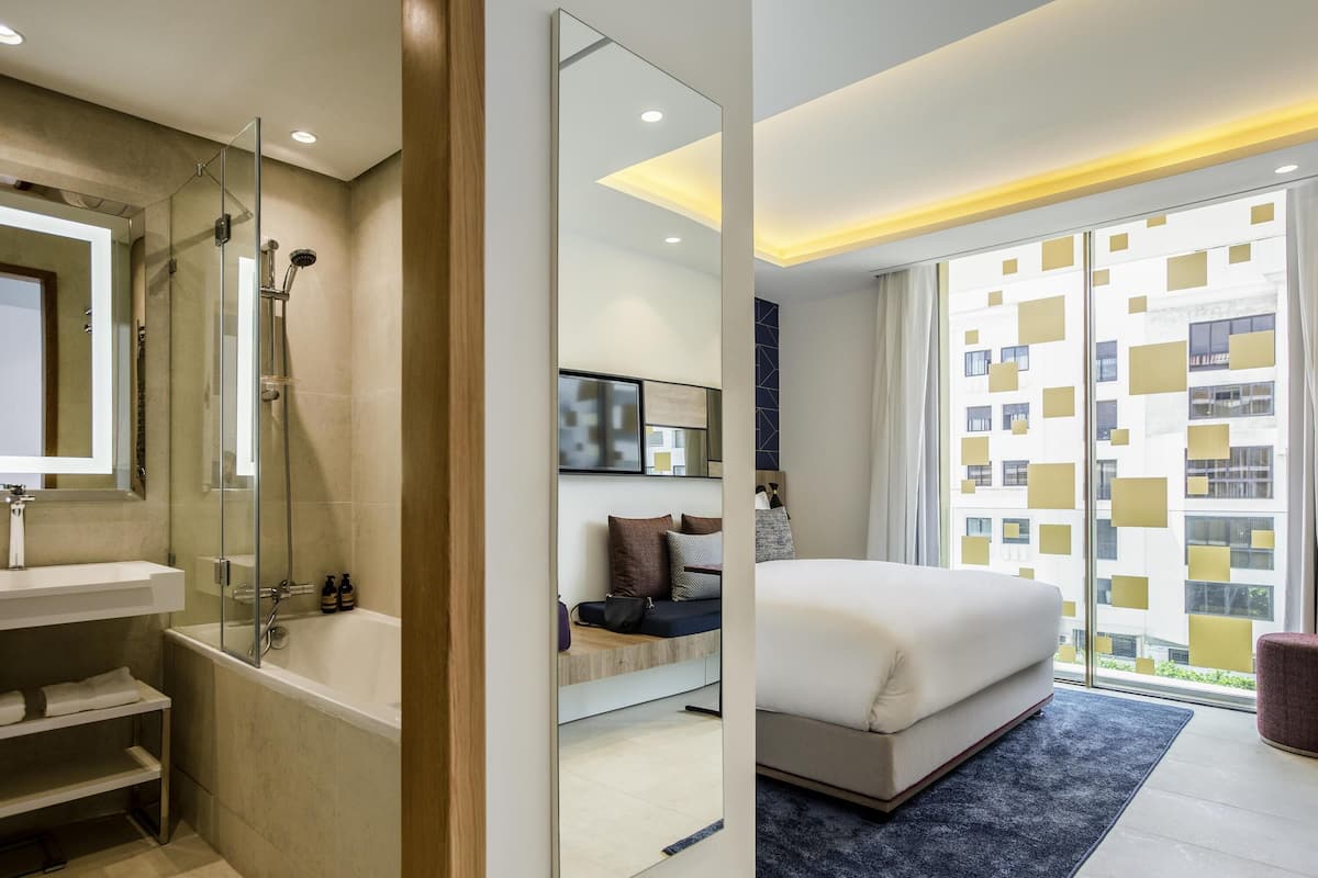 A modern hotel room with a large bed, a separate bathroom with a bathtub, a large mirror, and a view of the city through the window.