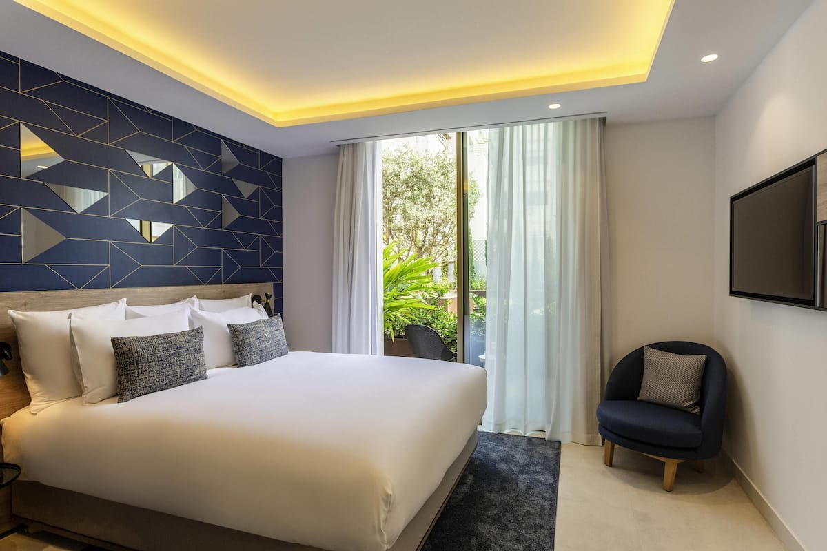 A modern hotel room with a large bed, a seating area, and a view of greenery outside.