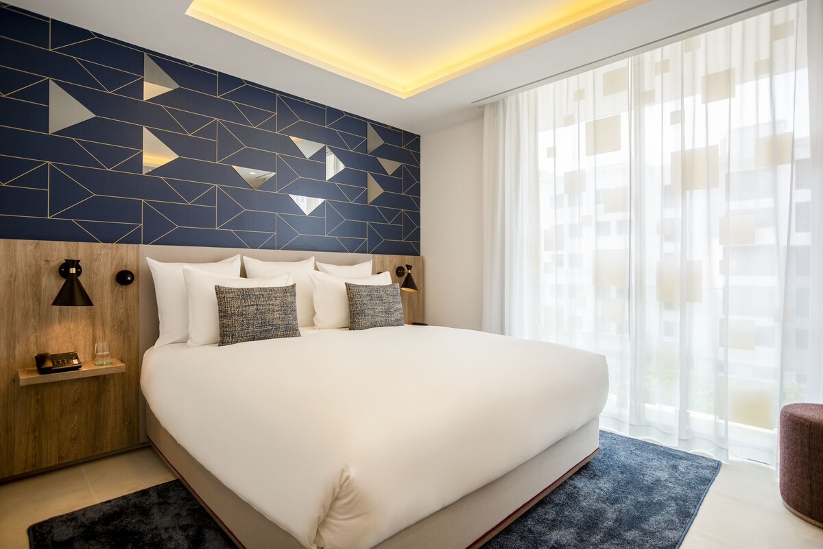 A large bed with white linens, a geometric-patterned wall, and a large window with sheer curtains.