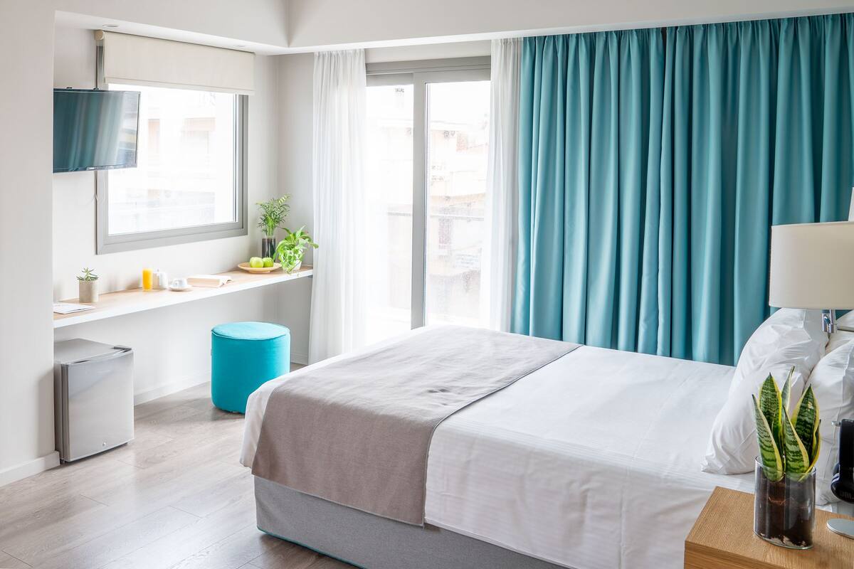 A hotel room with a bed, a TV mounted on the wall, a small desk with a plant, and a window with curtains.