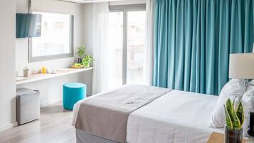 A hotel room with a bed, a TV mounted on the wall, a small desk with a plant, and a window with curtains.