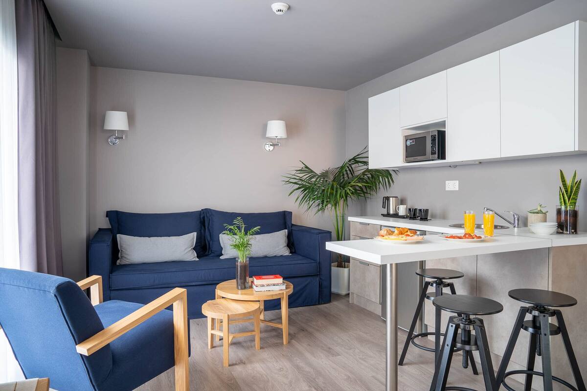 A modern living room with a blue sofa, a small wooden table with books, and a kitchen area with a microwave and bar stools.
