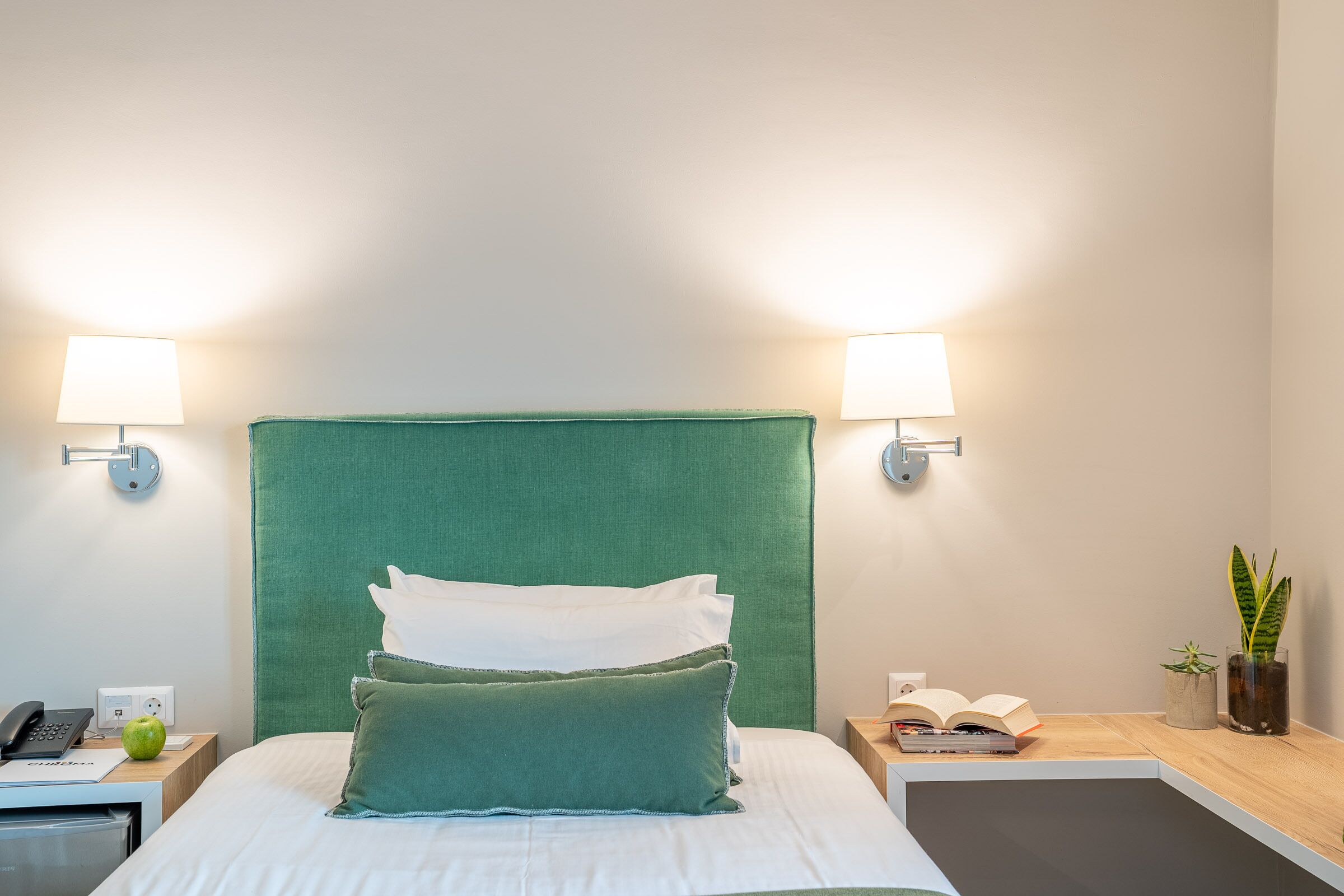 A neatly made bed with a green headboard, two bedside lamps, a bedside phone, a green apple, a book, and a small plant on a nightstand.