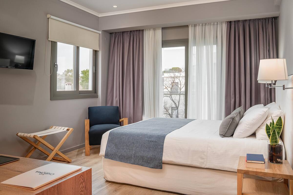 A modern hotel room with a large bed, a TV mounted on the wall, a chair, a small table with a lamp, and a window with curtains.